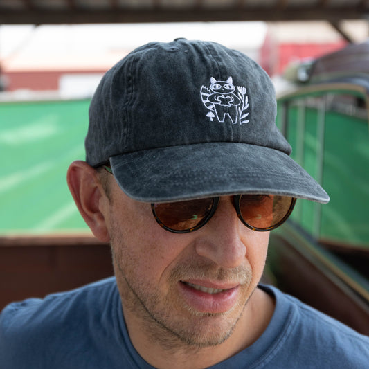 A man wearing a dark washed baseball cap with a white embroidered cartoon character logo, sunglasses, and a blue t-shirt, photographed outdoors under a sheltered structure.