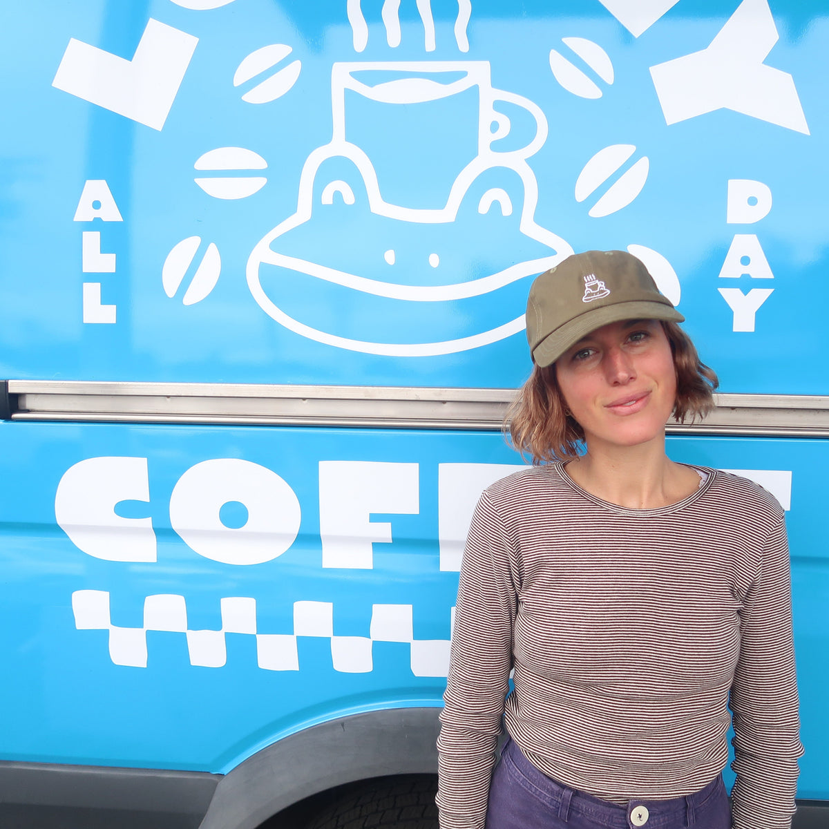 A woman wearing a brown long-sleeve shirt and a khaki baseball cap stands next to a bright blue vehicle with white coffee-themed graphics and the text 'COFFEE' visible.