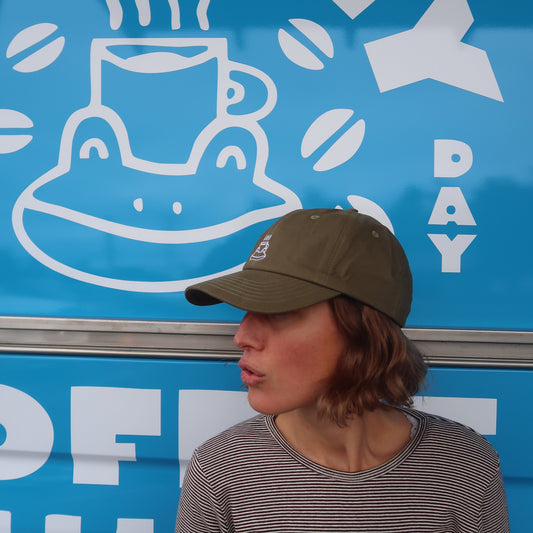 A woman wearing an olive green baseball cap and a gray striped long-sleeve shirt stands in front of a bright blue truck with white text reading 'COFFEE DAY' and a coffee cup graphic.