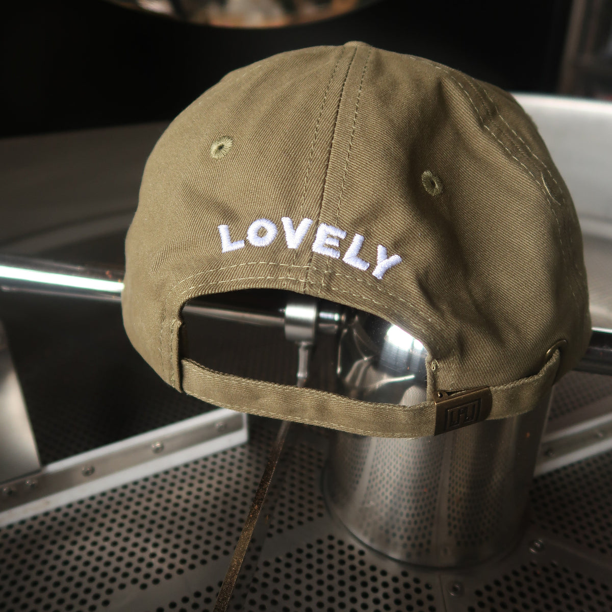 An olive green baseball cap with 'LOVELY' embroidered in white on the front, displayed on a metal stand inside a coffee shop setting.
