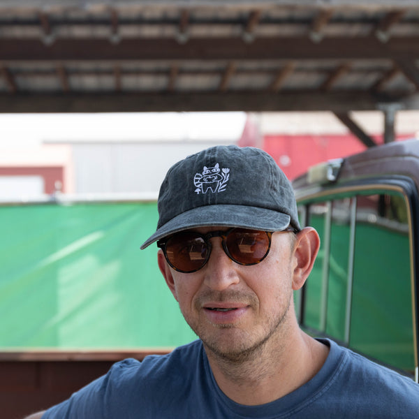 A man wearing a dark gray baseball cap with a white embroidered logo, dark sunglasses, and a blue t-shirt, standing in front of a green fence and a truck bed.