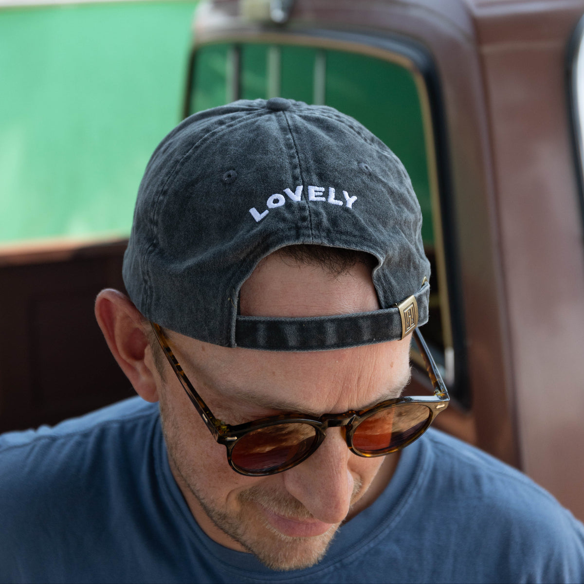 A man wearing a dark gray washed baseball cap with 'LOVELY' embroidered in white on the front, along with tortoiseshell sunglasses and a blue t-shirt, looking down in an outdoor setting.